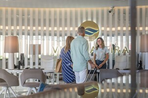 En moderne hotellobby med reception, siddeområde og et stort vindue med persienner.