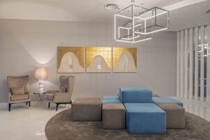 A modern interior with a seating area featuring a round rug, a coffee table, and two armchairs. There is a wall-mounted artwork and a geometric light fixture above the seating.