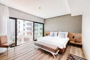 A modern hotel room with a large bed, a bench, a chair, a bedside table with a lamp, and a view of the city through a large window.