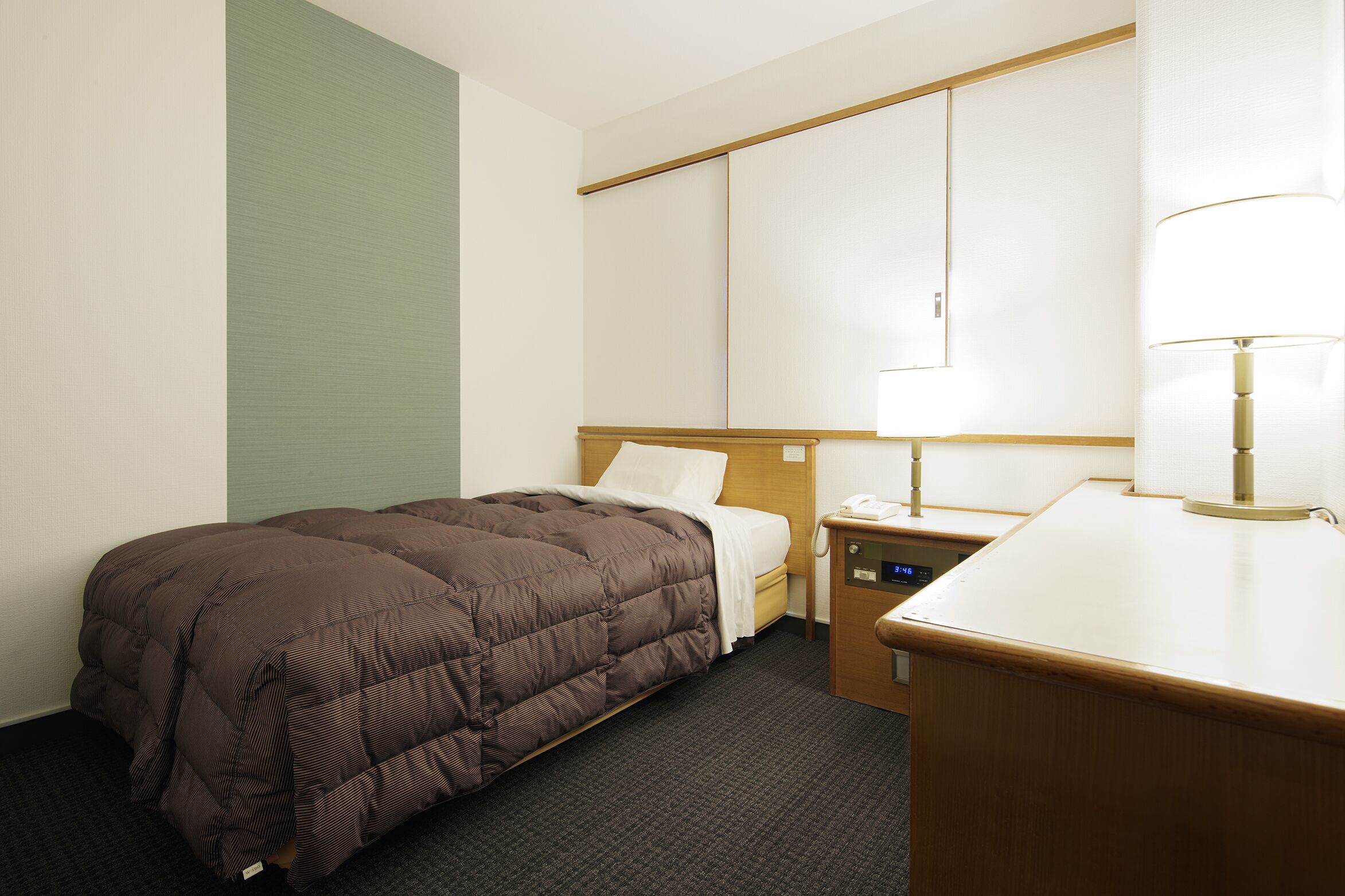 A hotel room with a single bed, a bedside table with a lamp, a small desk, and a wall-mounted TV.