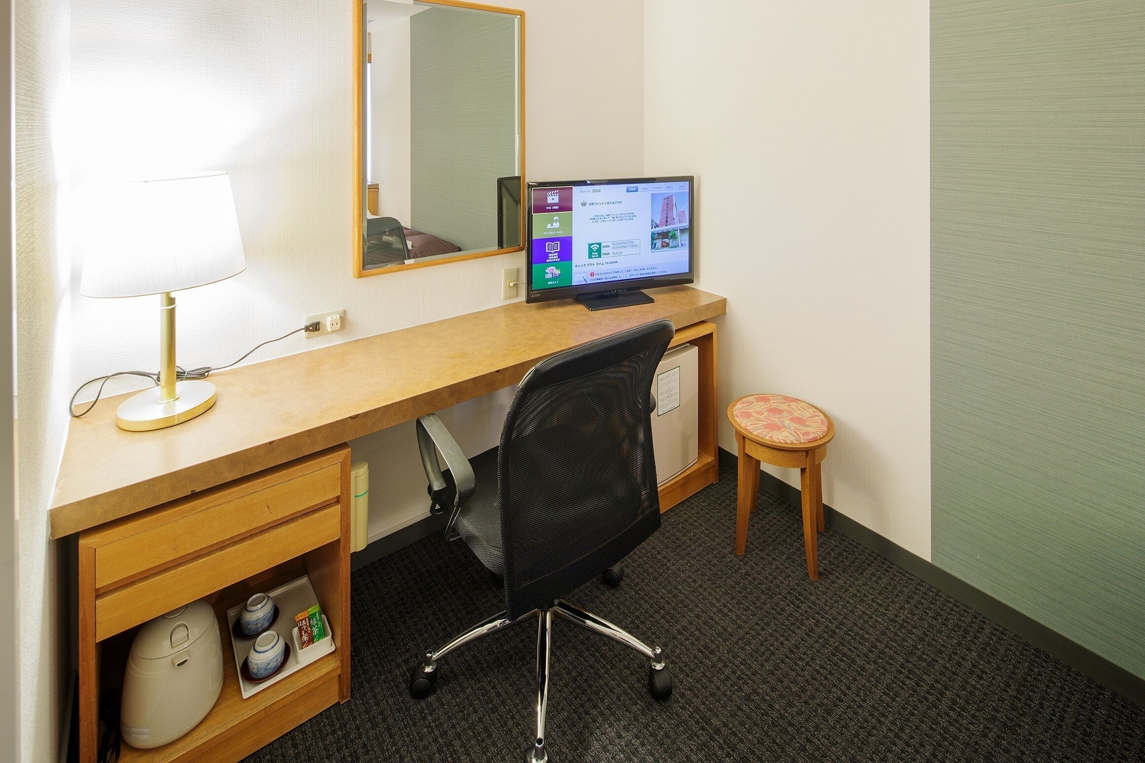 Habitación de hotel con un escritorio de madera, una computadora, una silla, un taburete pequeño y una tetera.