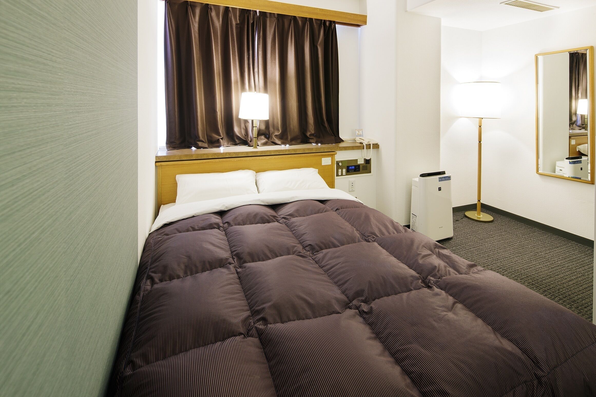 A hotel room with a bed, a bedside lamp, a mirror, and a wall-mounted heater.