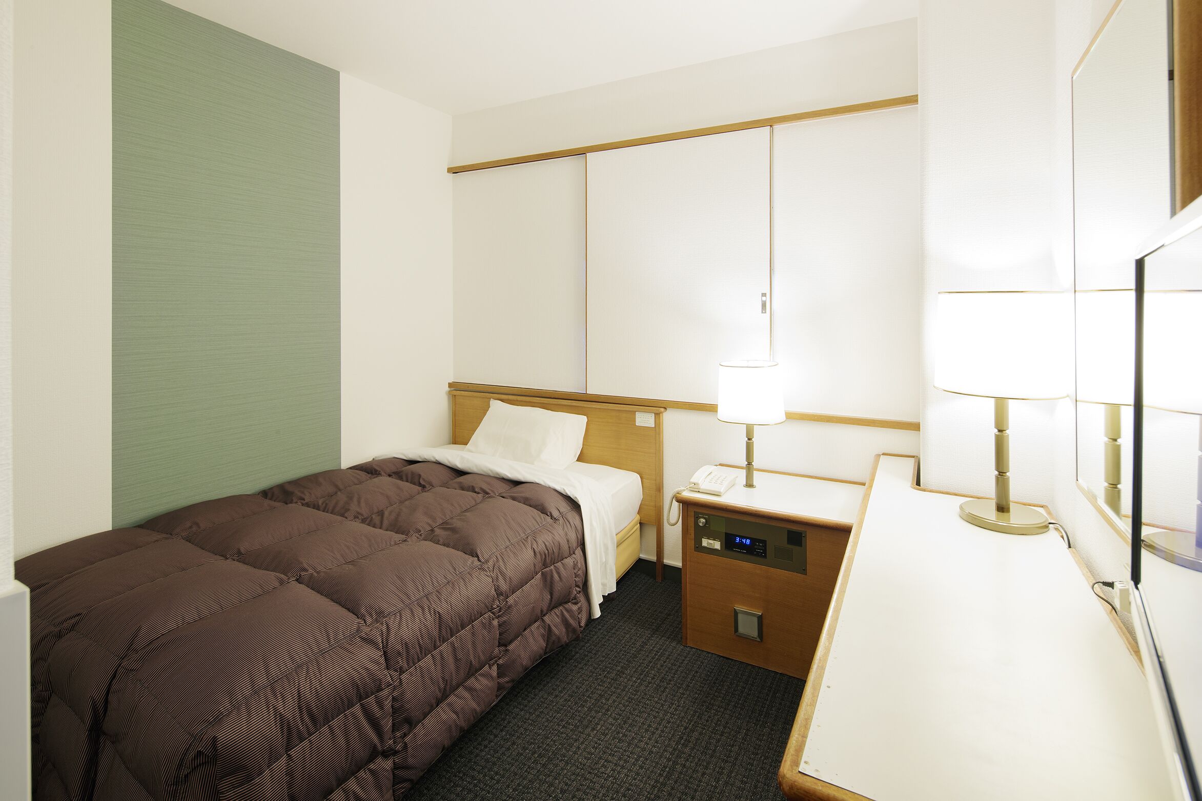 A hotel room with a bed, bedside table, lamp, and a small desk with a phone.