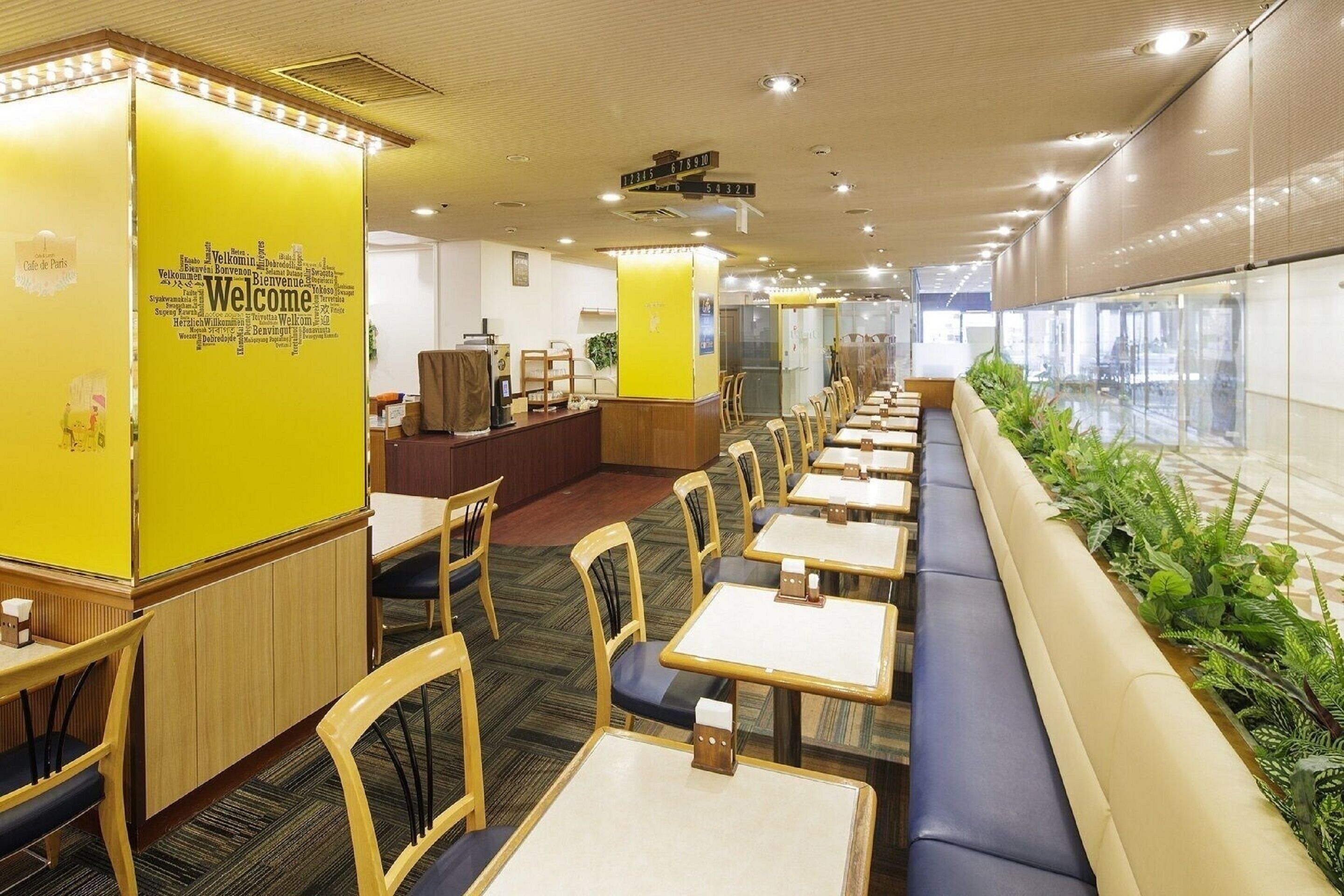 A modern indoor space with rows of tables and chairs, a reception area with a yellow wall, and a view through a glass wall.