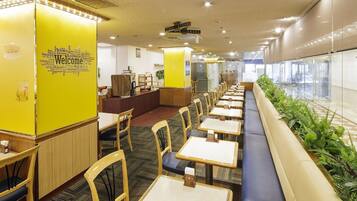 A modern indoor space with rows of tables and chairs, a reception area with a yellow wall, and a view through a glass wall.