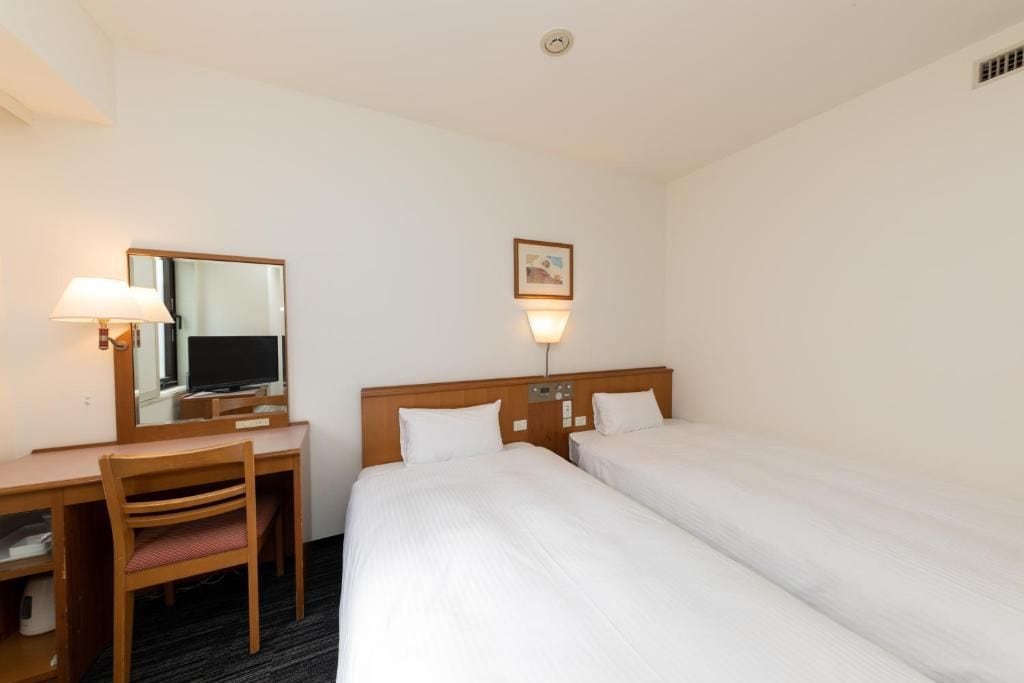 A hotel room with a desk, two beds, a chair, a mirror, a wall-mounted light, and a picture.