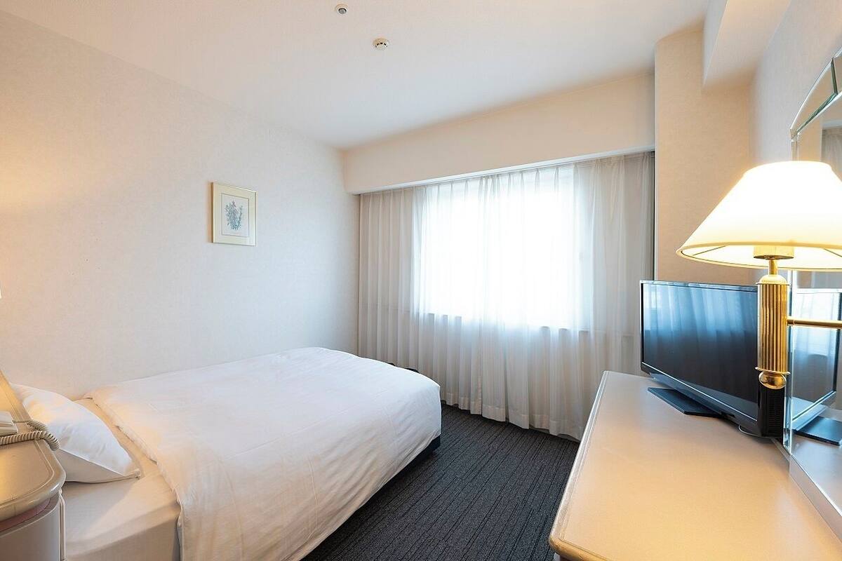 Une chambre d’hôtel avec un lit, un bureau doté d’une lampe, un téléphone, une télévision et une fenêtre avec des rideaux.