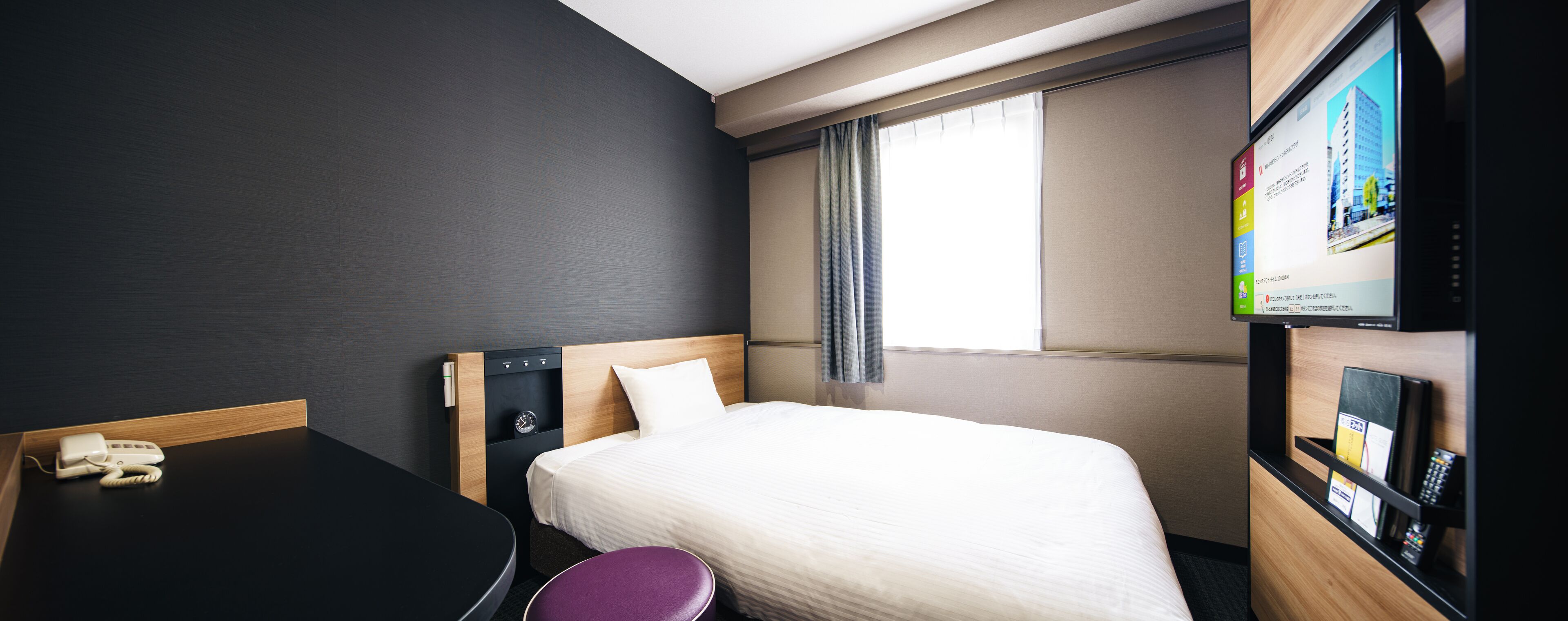 A hotel room with a bed, a desk, a chair, a phone, a clock, and a TV screen displaying a cityscape.