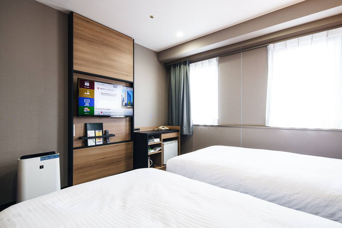 A hotel room with two beds, a desk with a computer, a small fridge, and a TV mounted on the wall.
