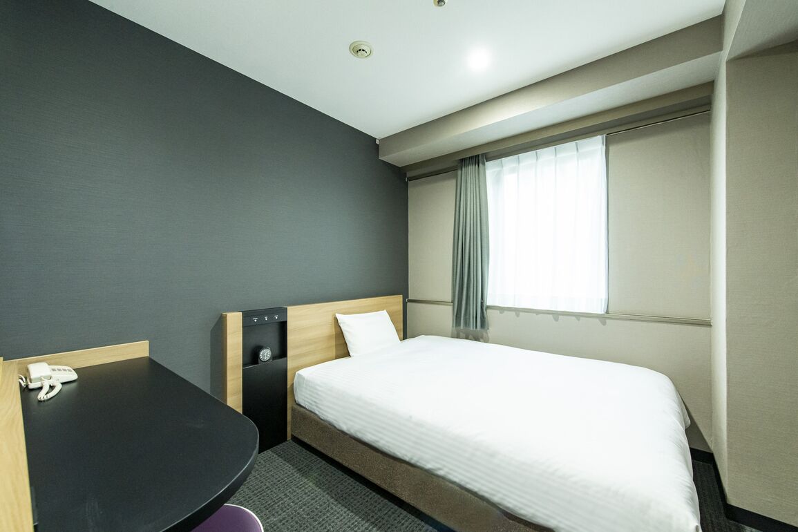 Una camera d'albergo con un letto, una scrivania, un telefono, una finestra con tende e una lampada a soffitto.