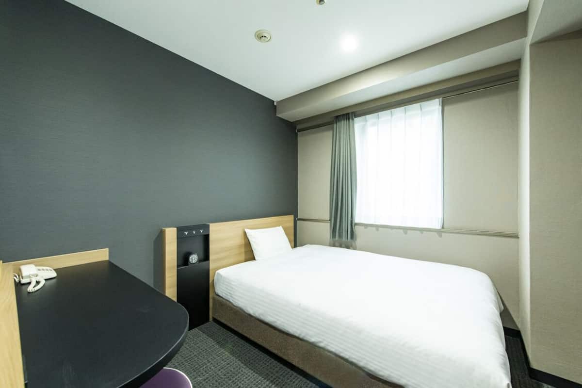 Habitación de hotel con cama, escritorio, teléfono, ventana con cortinas e iluminación en el techo.