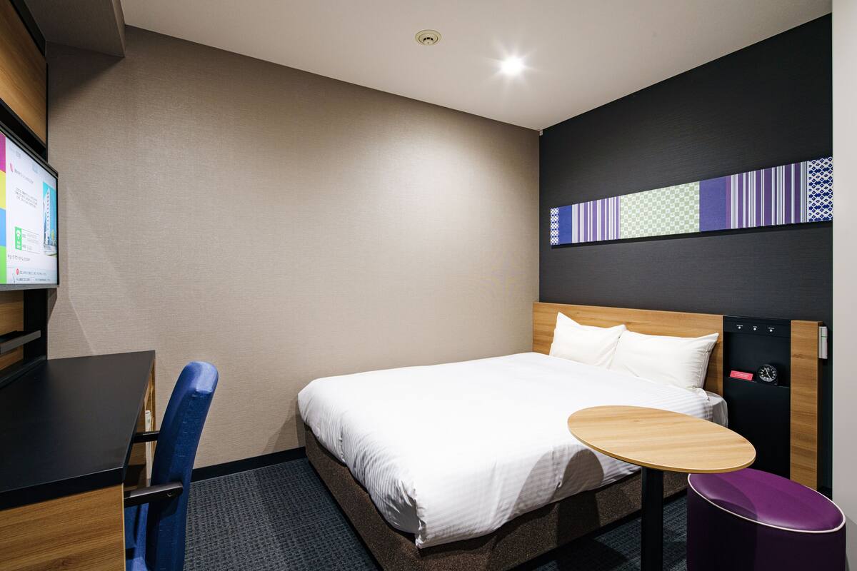 A hotel room with a bed, a desk, a chair, a small table, and a wall-mounted TV.