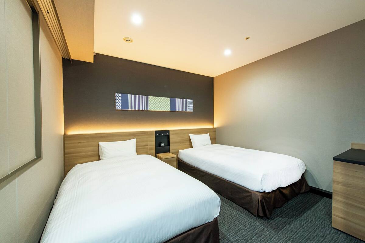 A hotel room with two beds, a small desk, a window with a patterned view, and a headboard with a light panel.