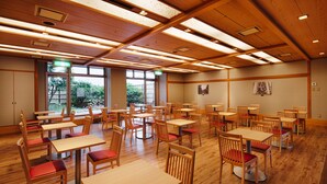 A spacious dining area with multiple wooden tables and chairs, large windows providing natural light, and a view of greenery outside.