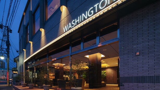 A hotel entrance with a large sign reading "WASHINGTON," outdoor seating with potted plants, and a brick facade.