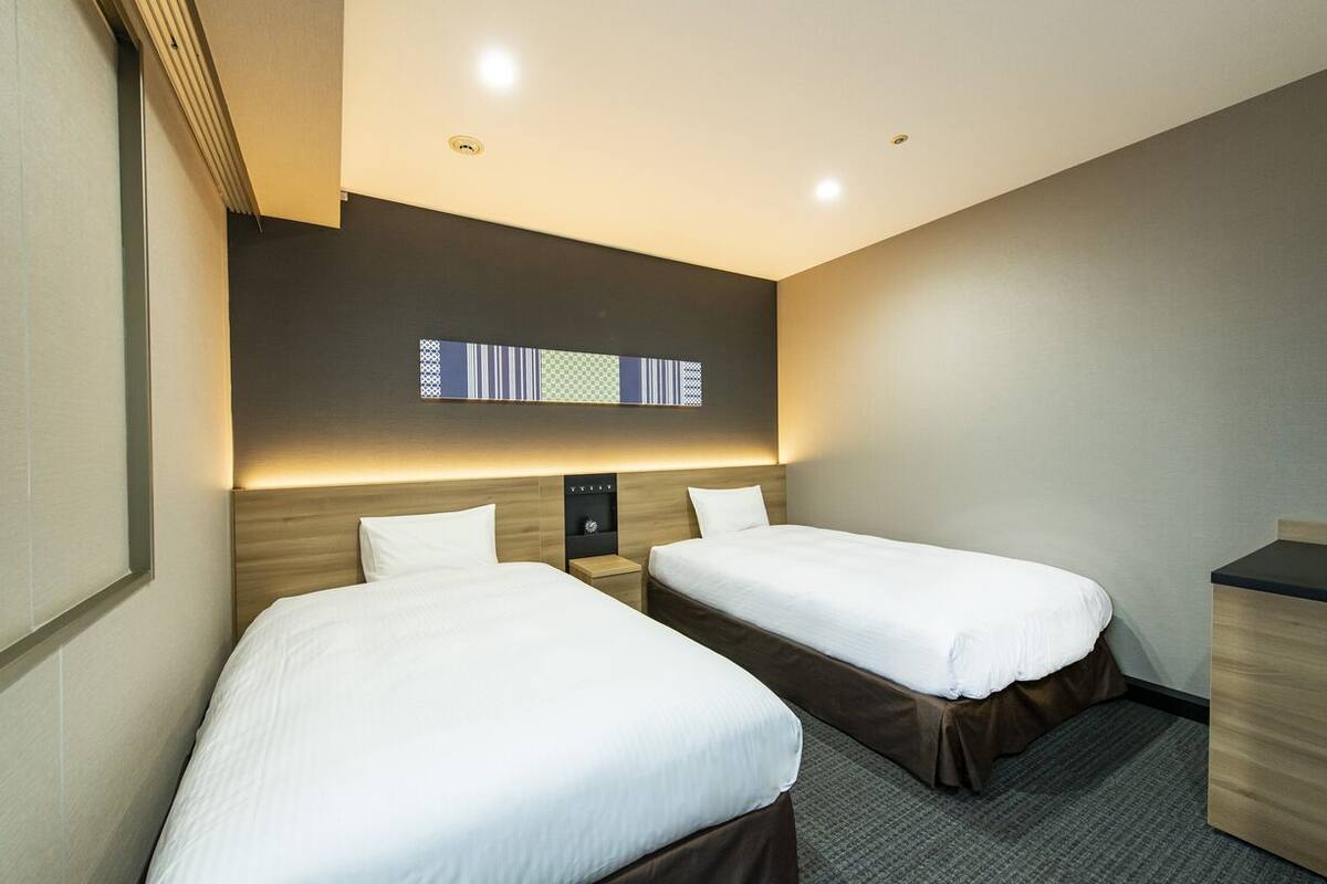 A hotel room with two beds, a small desk, a window with patterned blinds, and a wall-mounted light fixture.