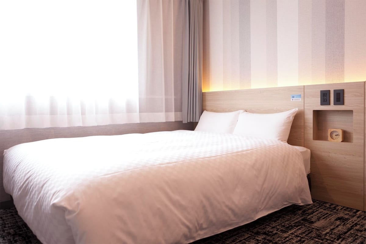 Ein Hotelzimmer mit einem großen Bett, einem hölzernen Kopfteil, einem Nachttisch mit einer Uhr und durchsichtigen Vorhängen.