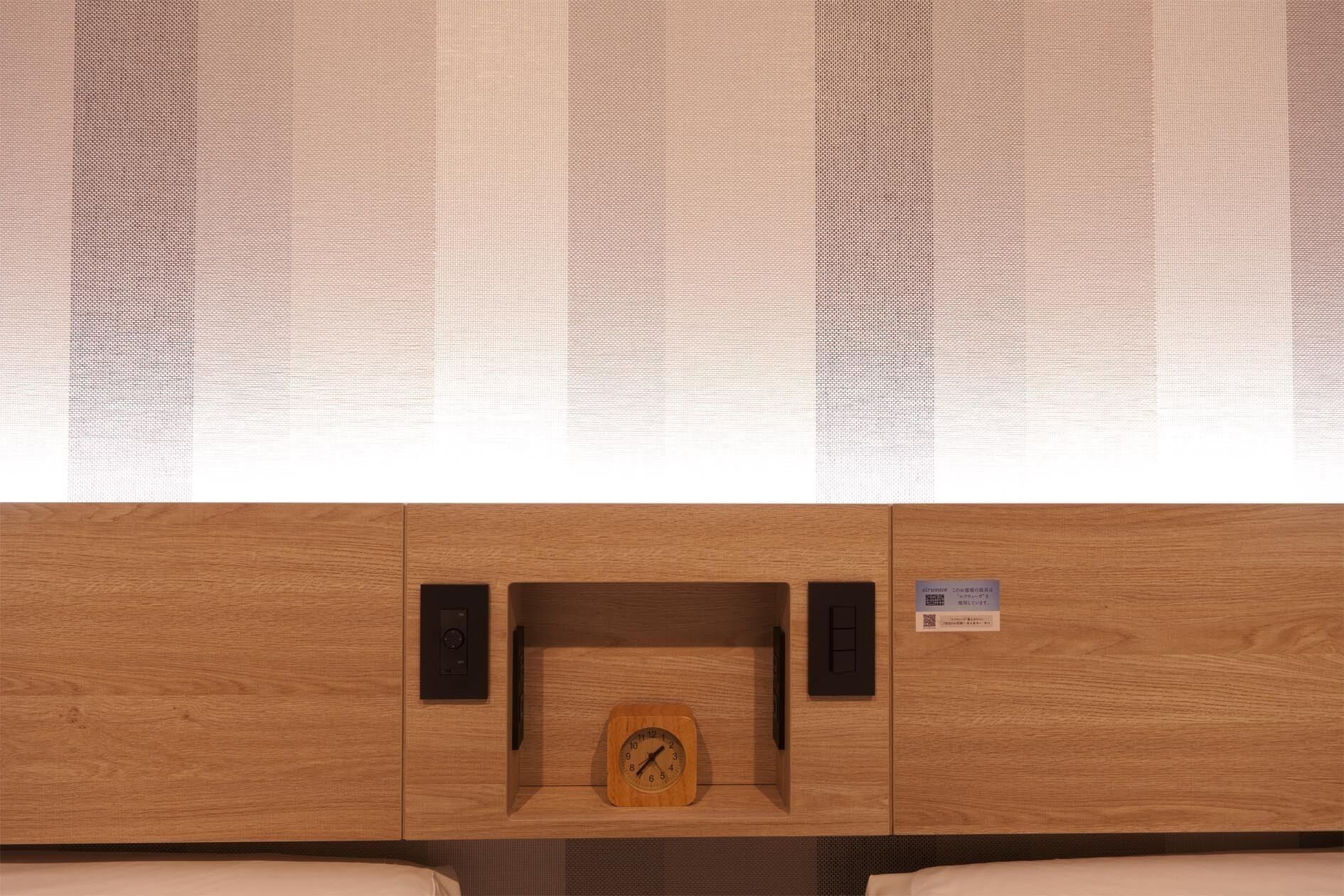 Un mueble de madera con un reloj y un estante pequeño, una ventana con una persiana decorativa, y una cama con un colchón blanco.