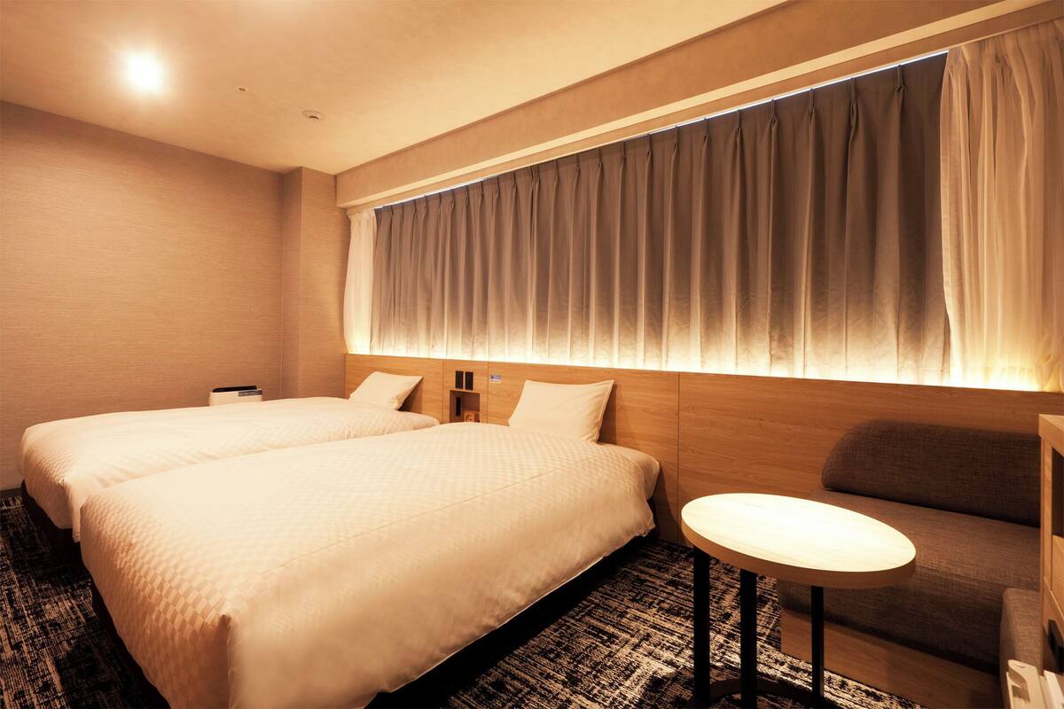 Ein Hotelzimmer mit zwei Betten, einem kleinen runden Tisch, einer Sitzecke und einem Fenster mit Vorhängen.