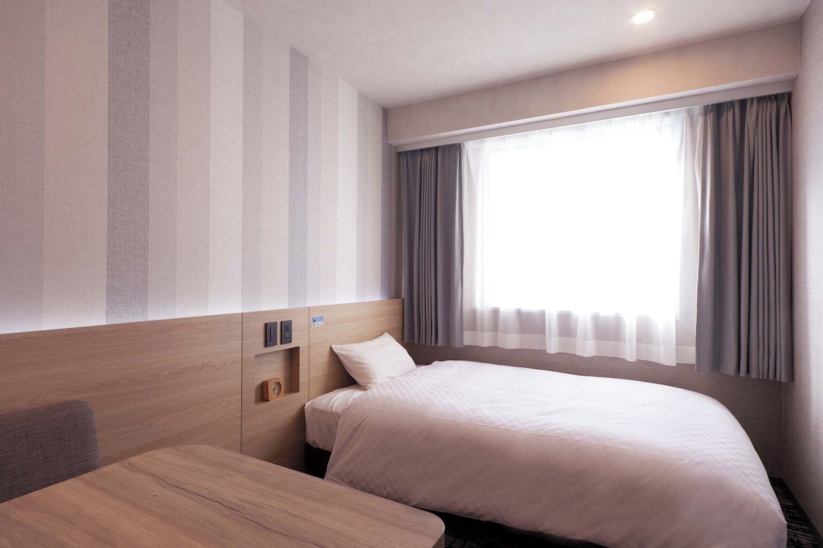 Ein Hotelzimmer mit einem Bett, einem Nachttisch aus Holz, einem kleinen Tisch und einem großen Fenster mit Vorhängen.