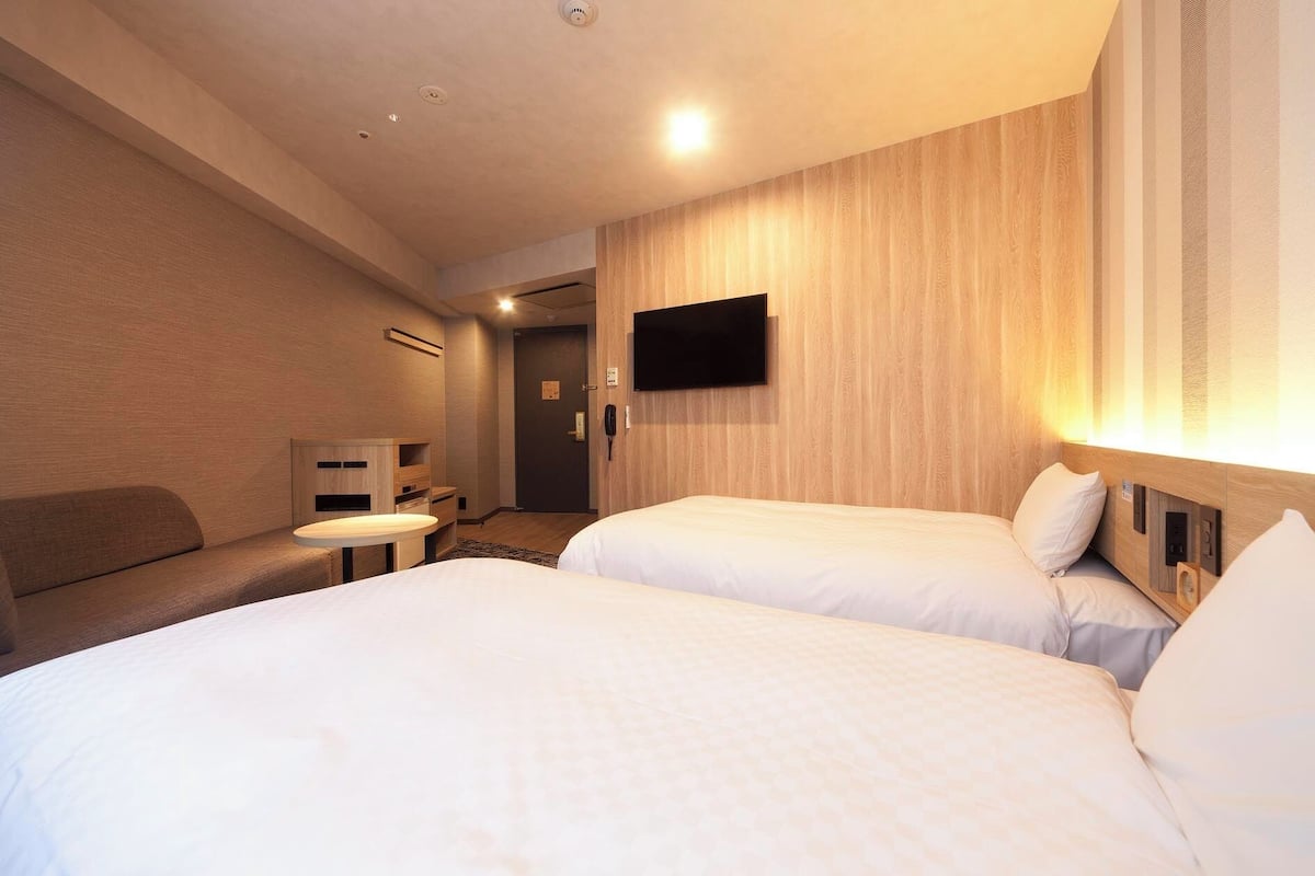 Ein Hotelzimmer mit zwei Betten, einem an der Wand befestigten Fernseher, einem Schreibtisch und einem Sofa.