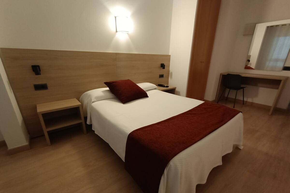 A hotel room with a wooden headboard, a single bed with white bedding and a red throw, a bedside table, a desk with a chair, and a wall-mounted light.
