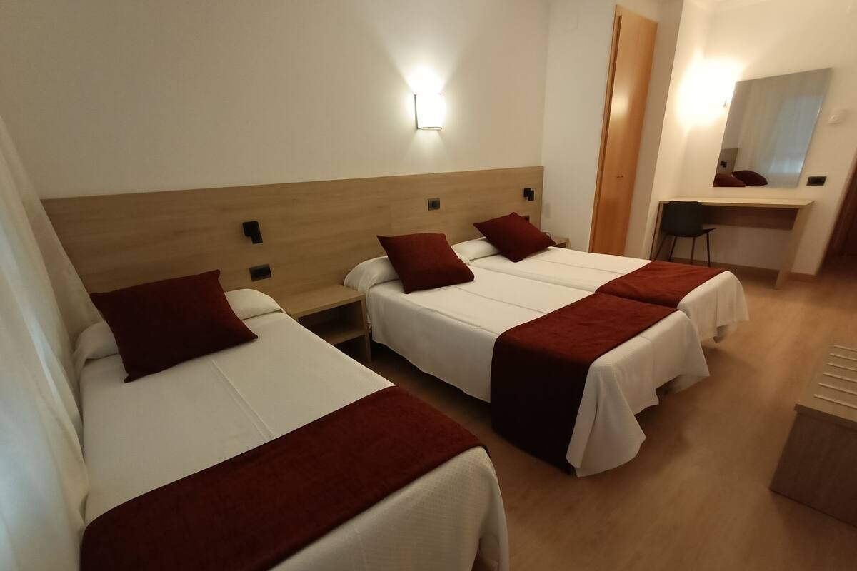A hotel room with two single beds, each with a red throw blanket and white bedding. There is a wooden headboard, a nightstand, and a desk with a chair. A wall-mounted light fixture is visible above the bed.