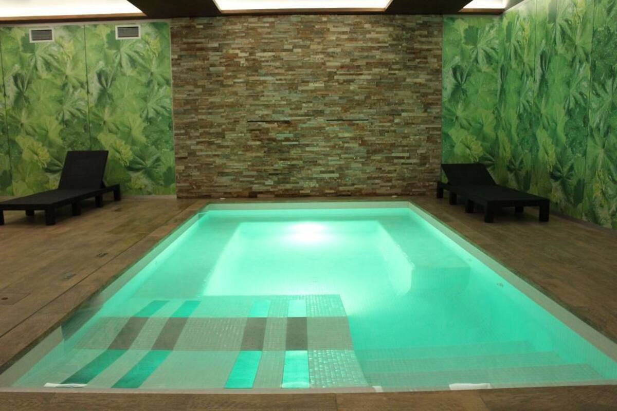 A rectangular indoor swimming pool with a tiled floor and a stone wall in the background.