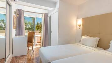 A hotel room with two beds, a sitting area with a chair, a table, and a view of palm trees and a pool.