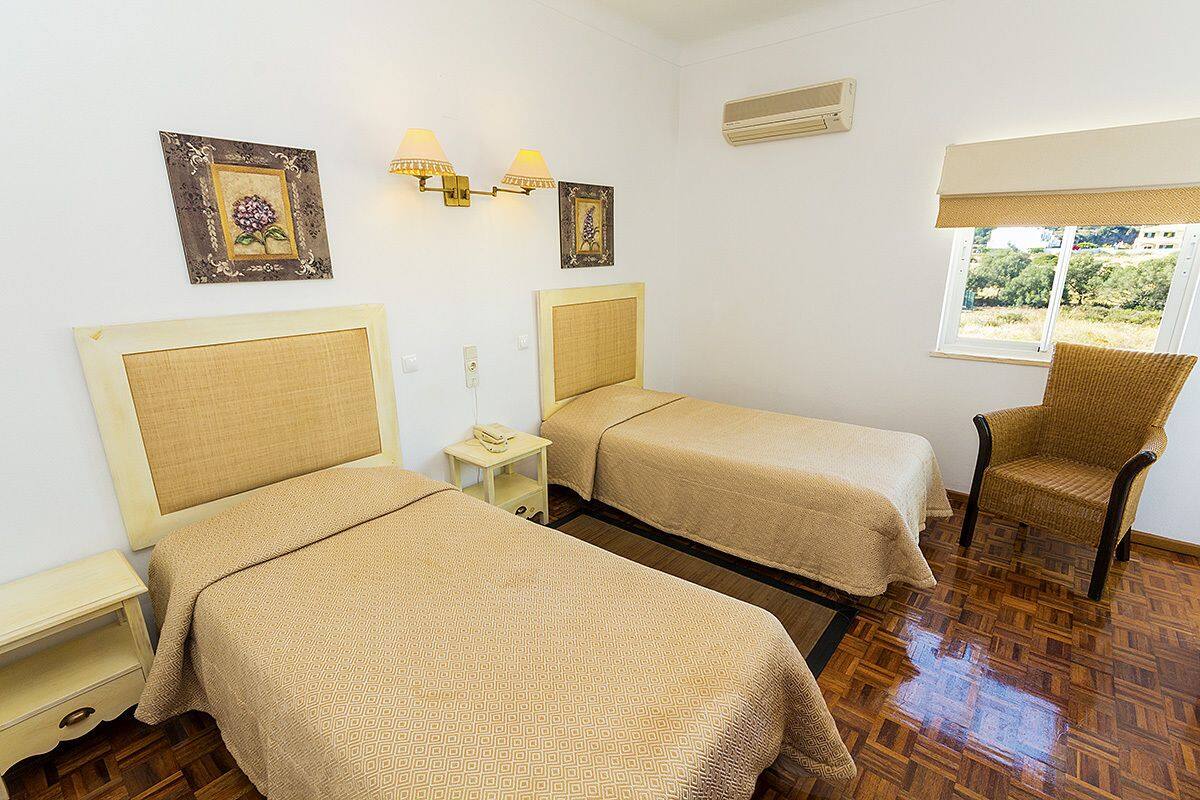 A room with two beds, a chair, a small table, wall-mounted lamps, and a window with a view.