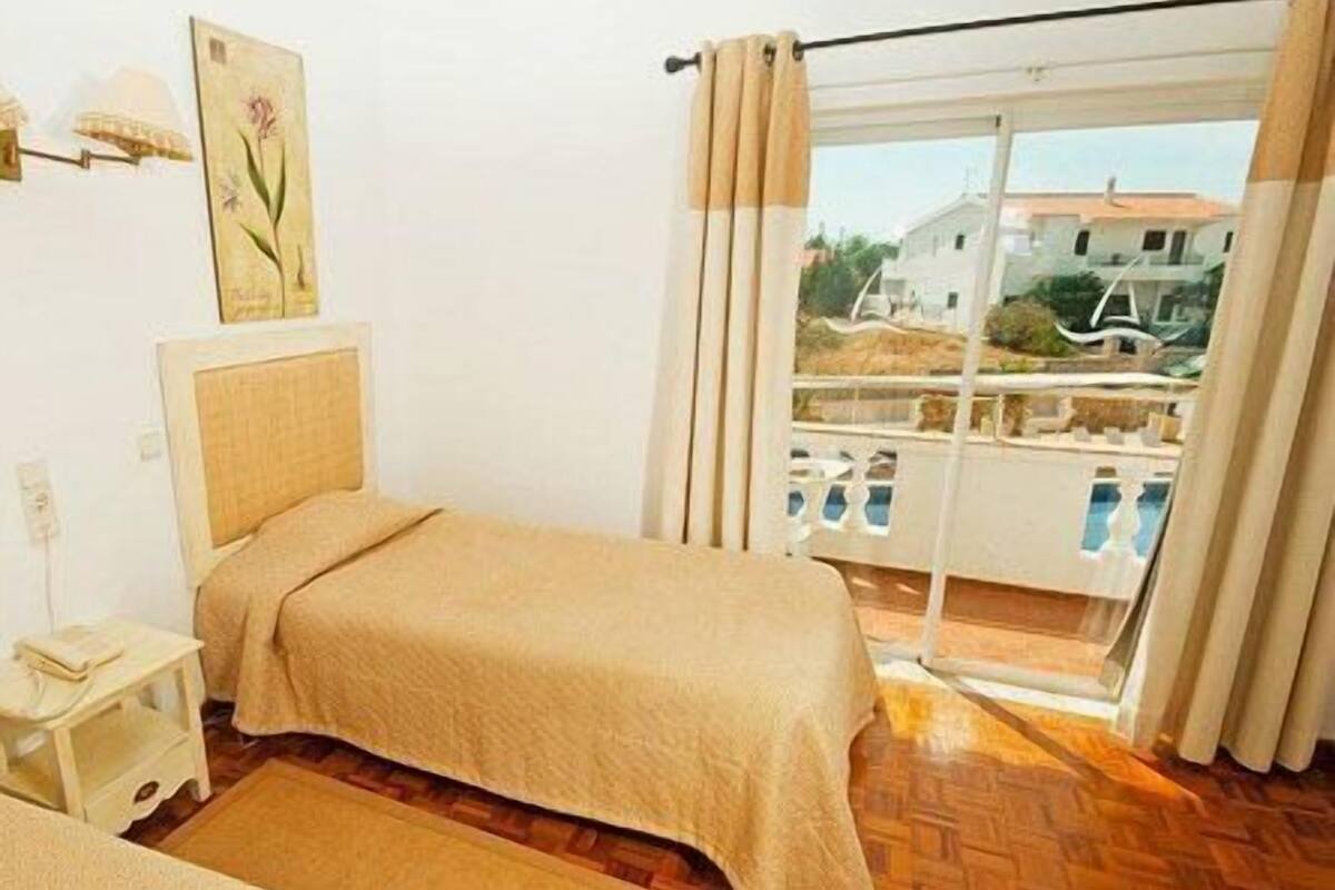 A bedroom with a bed, a nightstand, a painting, and a view of a balcony with a pool.