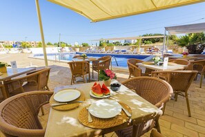 An outdoor dining area with wicker chairs and tables, a pool in the background, and a clear sky. - Vila Channa (Albufeira)