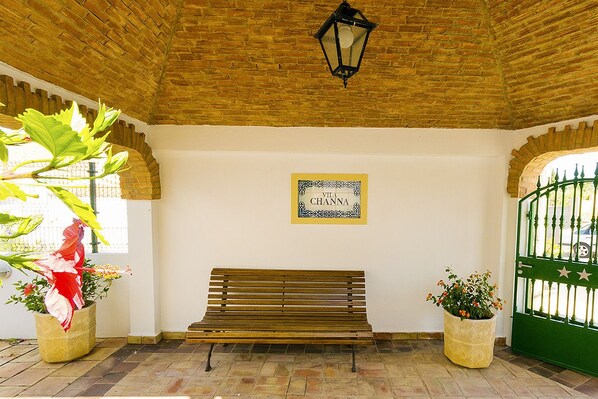 A bench in a covered area with a brick ceiling, a sign reading "Villa Channa," and two potted plants.
