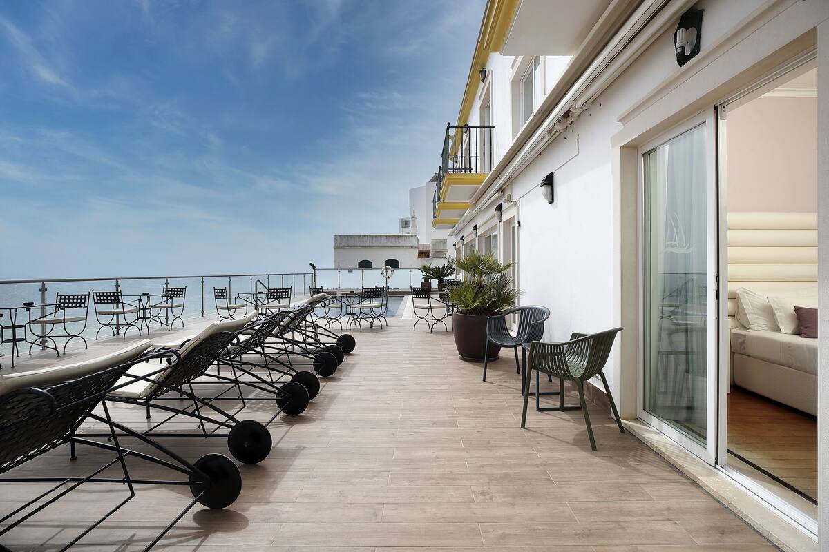 Une terrasse extérieure ensoleillée, dotée de chaises longues et de tables, d’une plante en pot et d’une porte-fenêtre coulissante donnant sur un espace intérieur.