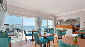 Een restaurant met blauwgroene stoelen en houten tafels, grote ramen met uitzicht op zee en een bar met flessen op de planken.