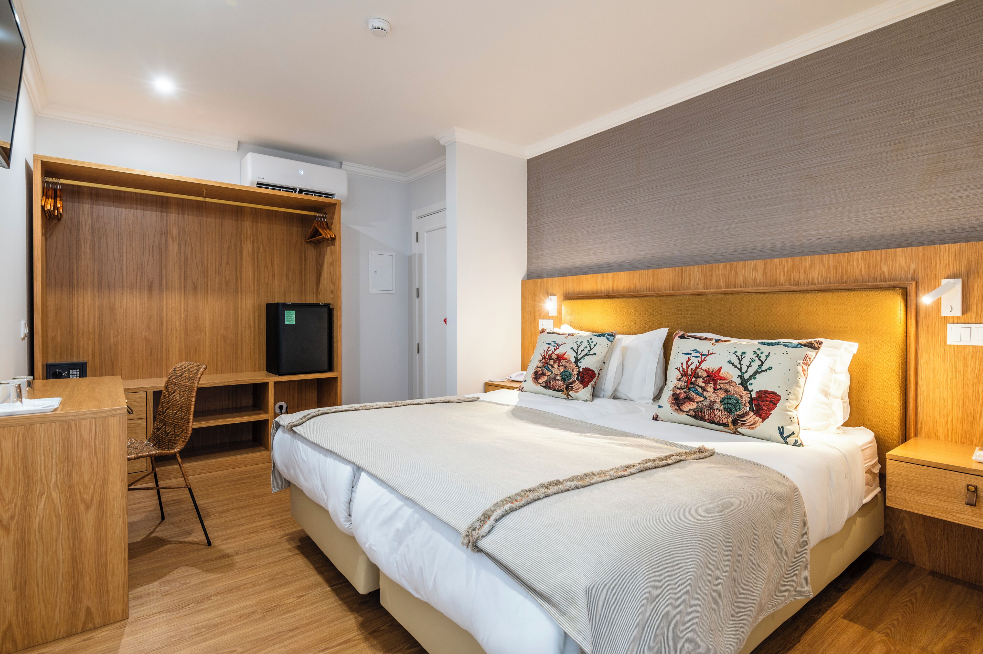 A hotel room with a bed, bedside table, desk, chair, wardrobe, and an air conditioning unit.