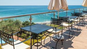 Een terras buiten met tafels en stoelen, met uitzicht op een heldere blauwe zee en de kust.
