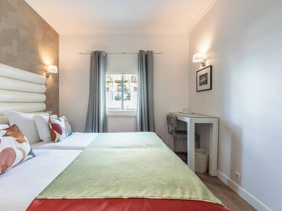 A hotel room with a bed, a desk, a chair, a window with curtains, and a wall-mounted lamp.