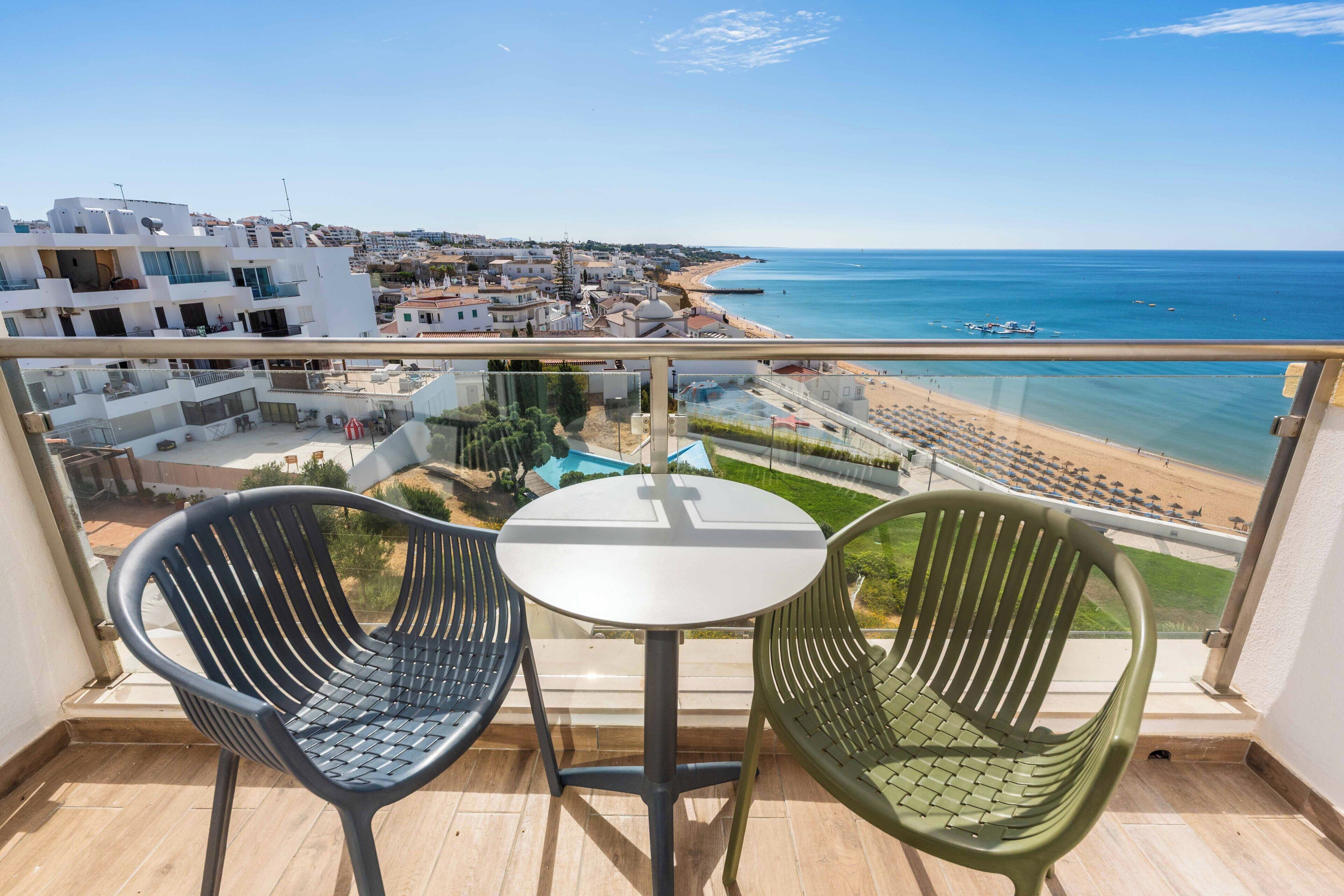 Un balcón con dos sillas y una mesa, con vista a una playa y un pueblo costero.