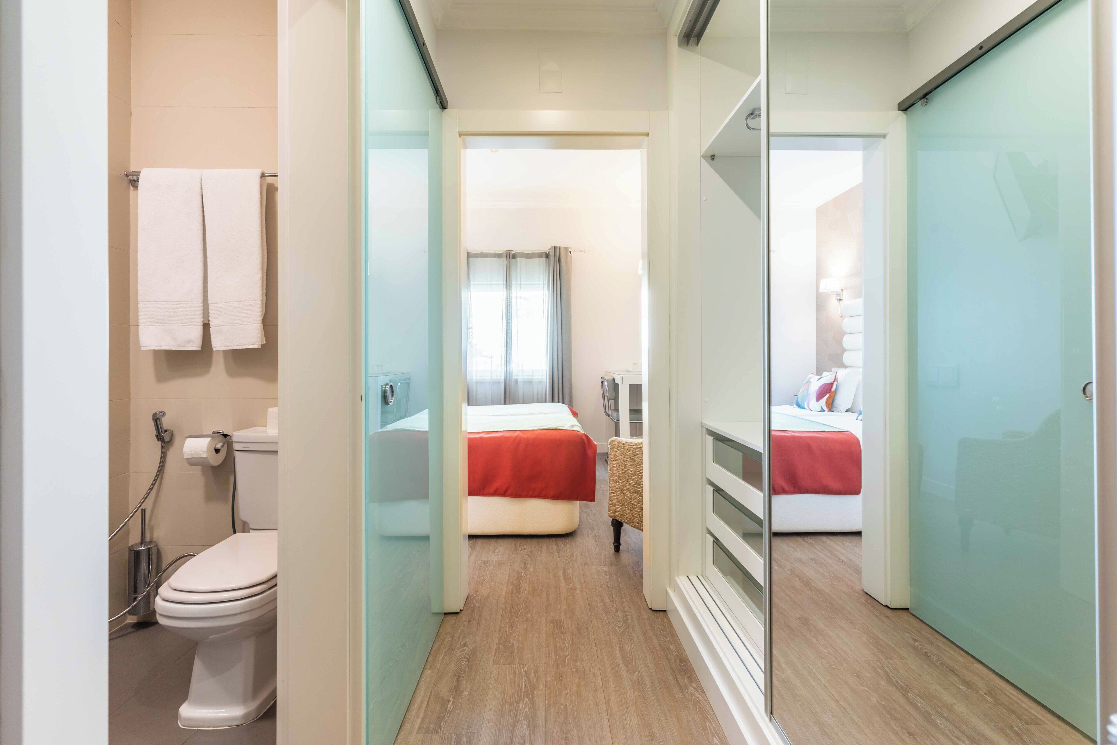 A hotel room with a toilet, a bed with a red blanket, a chair, and a glass partition.