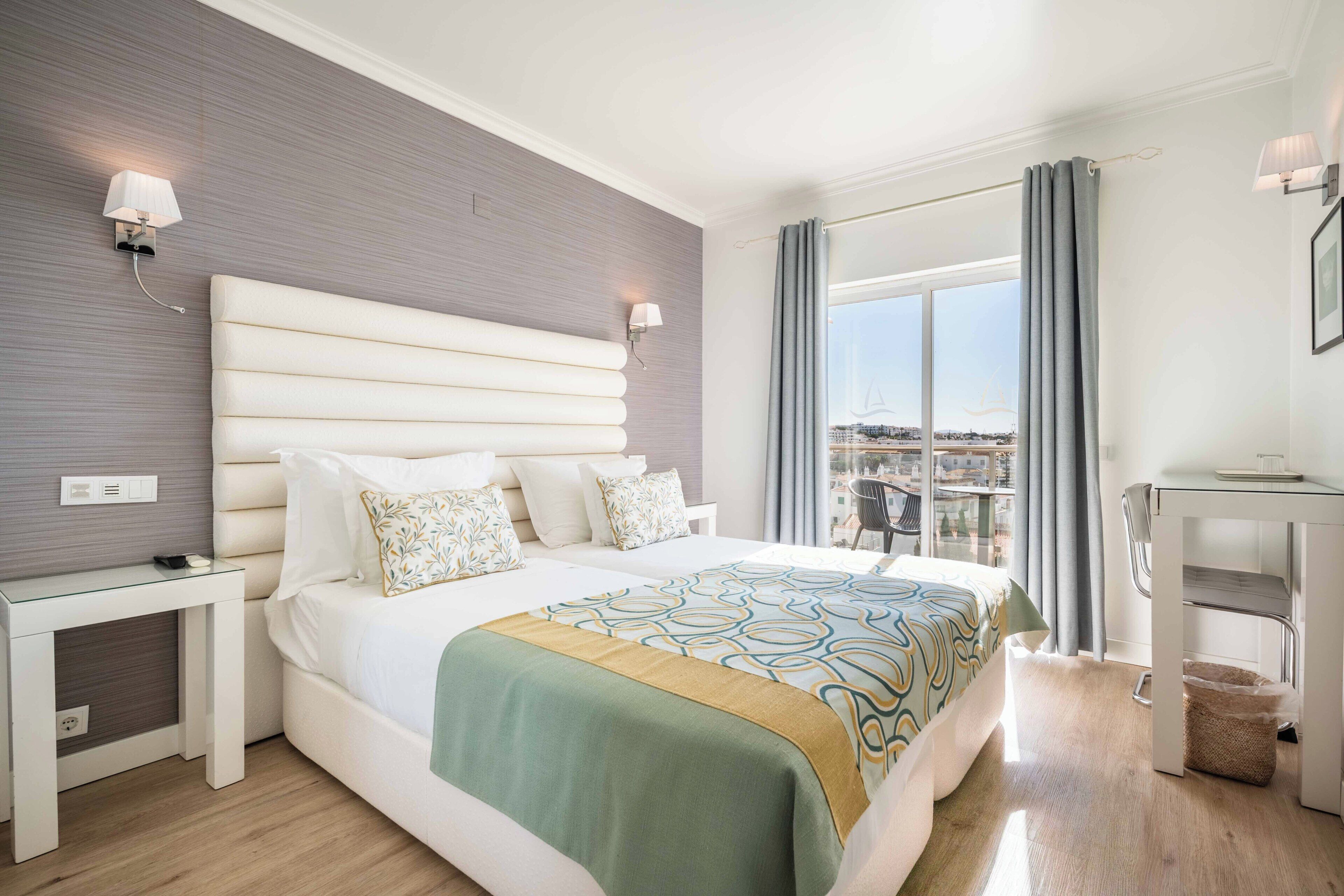 Habitación de hotel con una cama grande, una mesita de noche, un escritorio, una silla, un ventanal con cortinas y vistas a la ciudad.