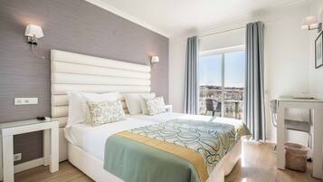 Habitación de hotel con una cama grande, una mesita de noche, un escritorio, una silla, un ventanal con cortinas y vistas a la ciudad.