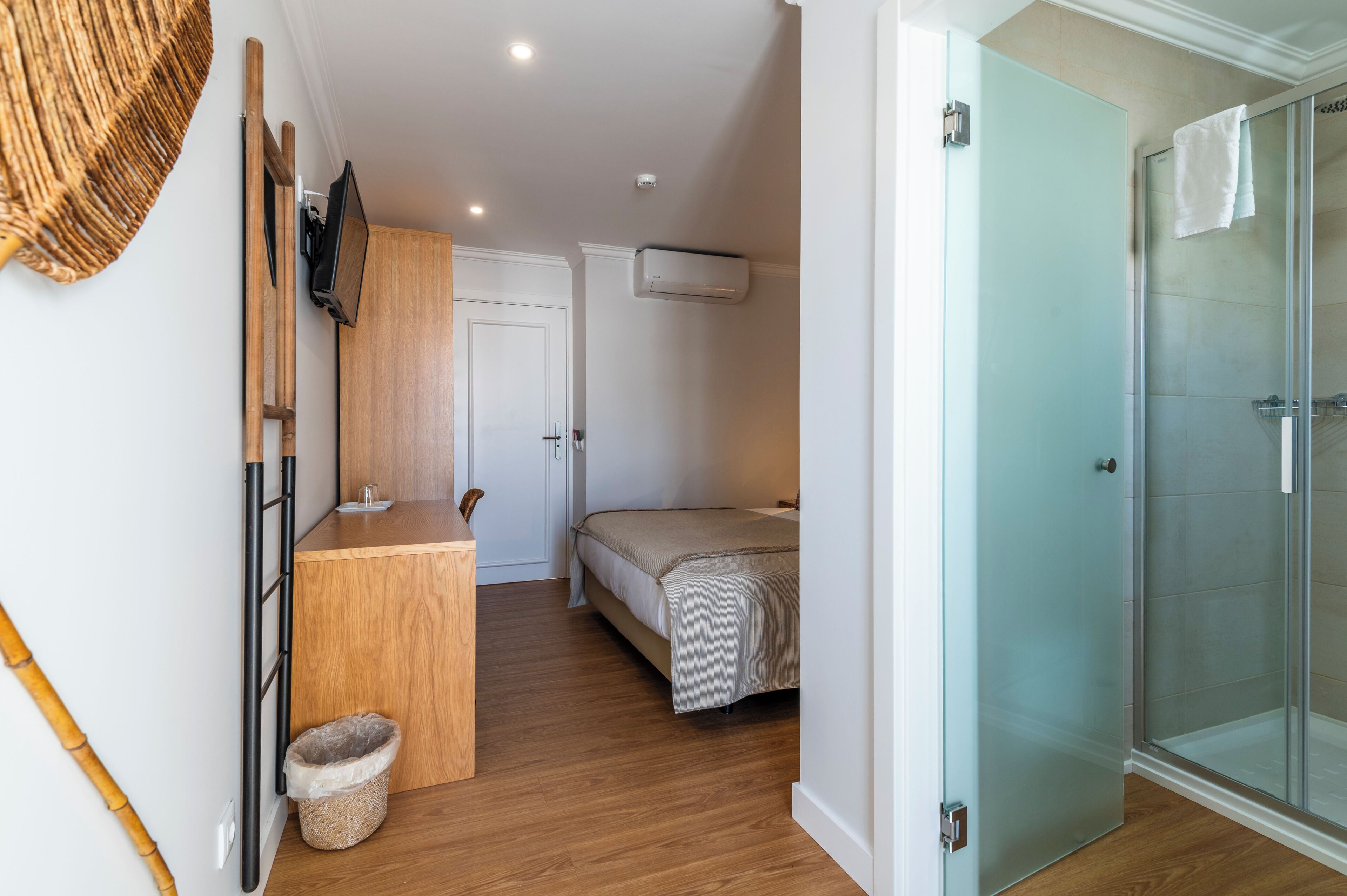 Un dormitorio compacto con cama, un escritorio de madera pequeño, un televisor montado en la pared y un box de ducha de vidrio.