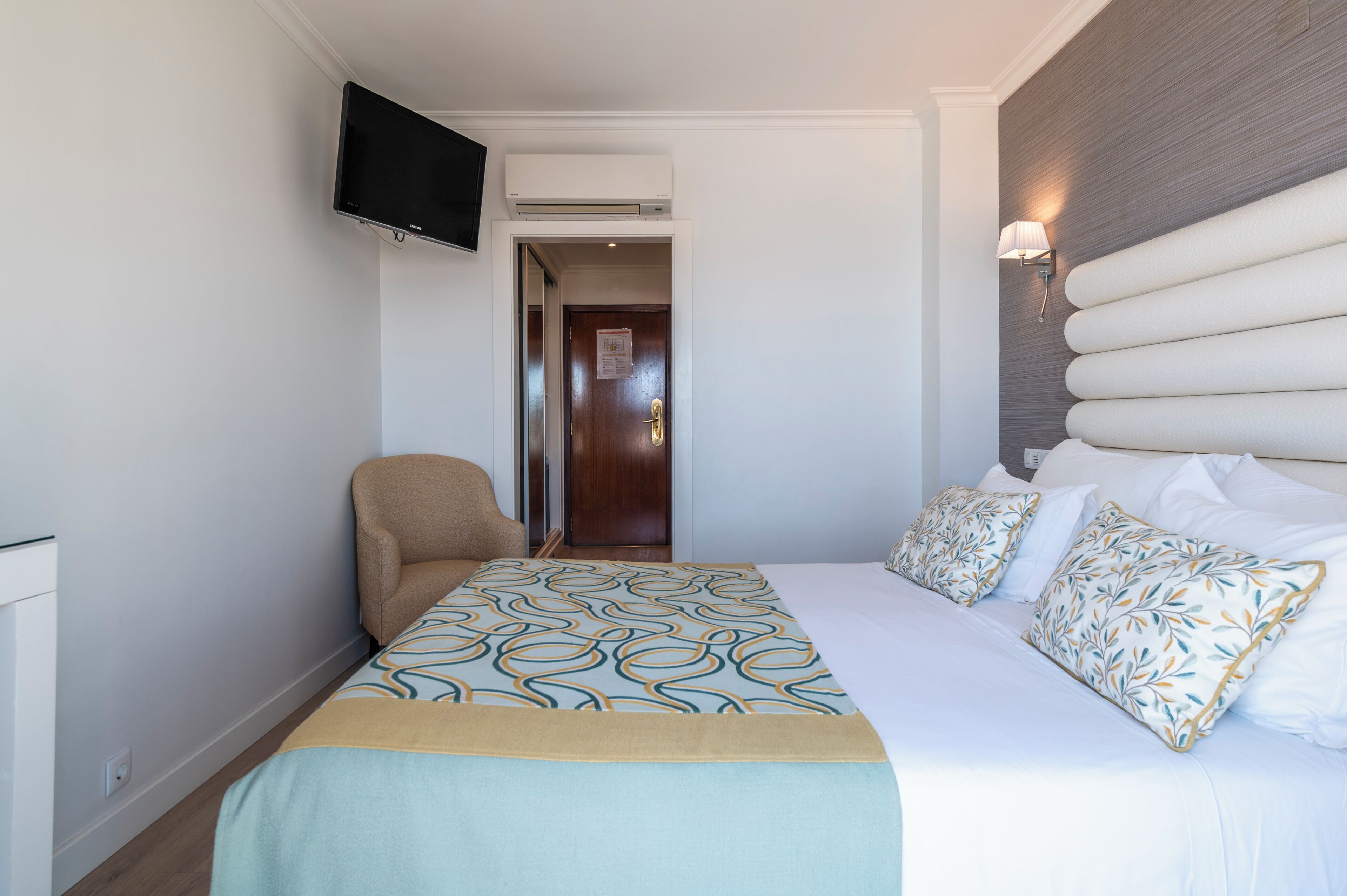Una habitación de hotel con cama, una silla, televisión y aire acondicionado instalado en la pared.