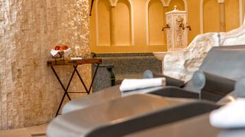 A spa room with a stone wall, a massage table, a small table with a towel and fruit, and a decorative cabinet.
