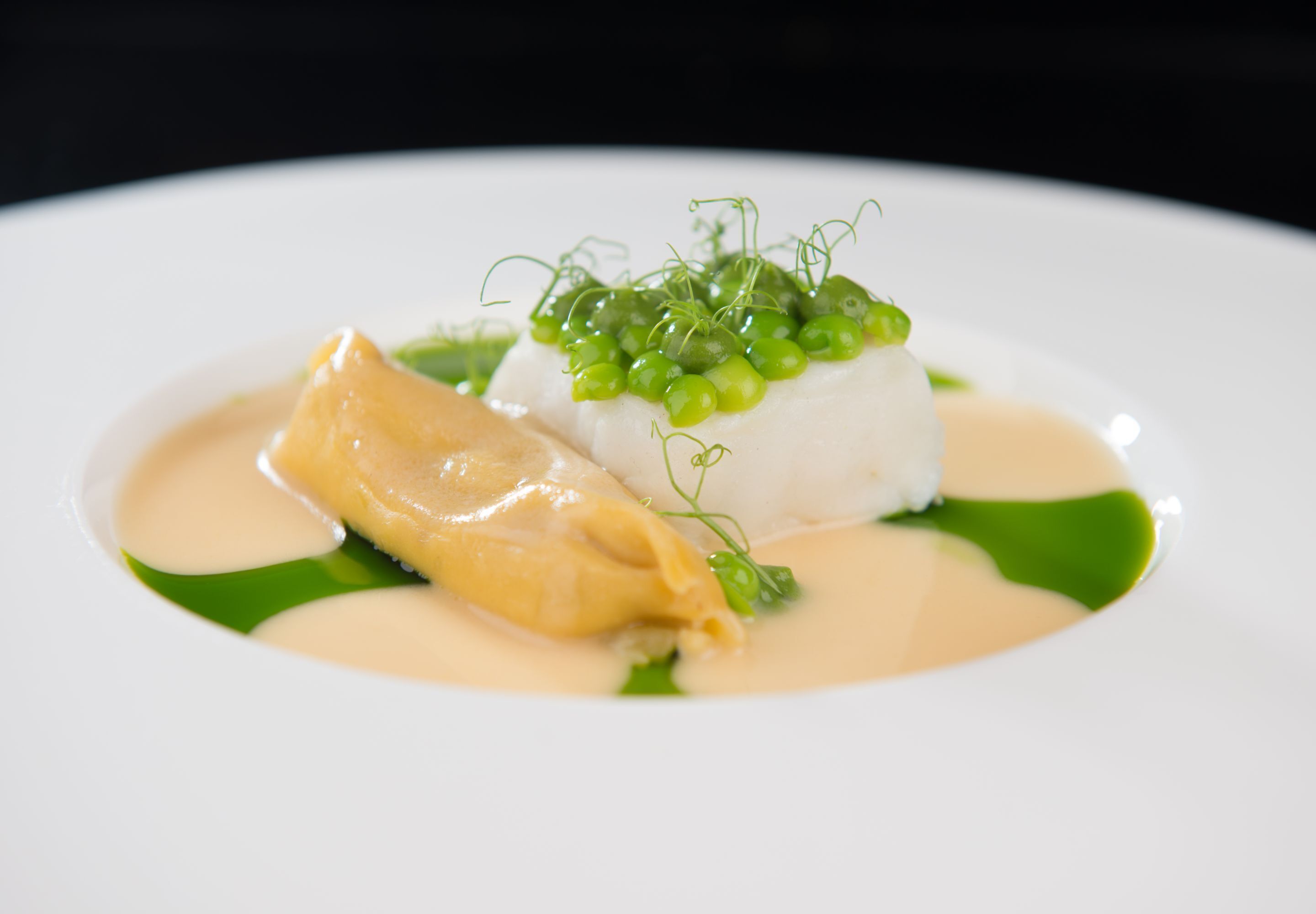 Ein weißer Teller mit einem Stück weißem Fisch, belegt mit grünen Erbsen und einem Stück gelbem Fisch.