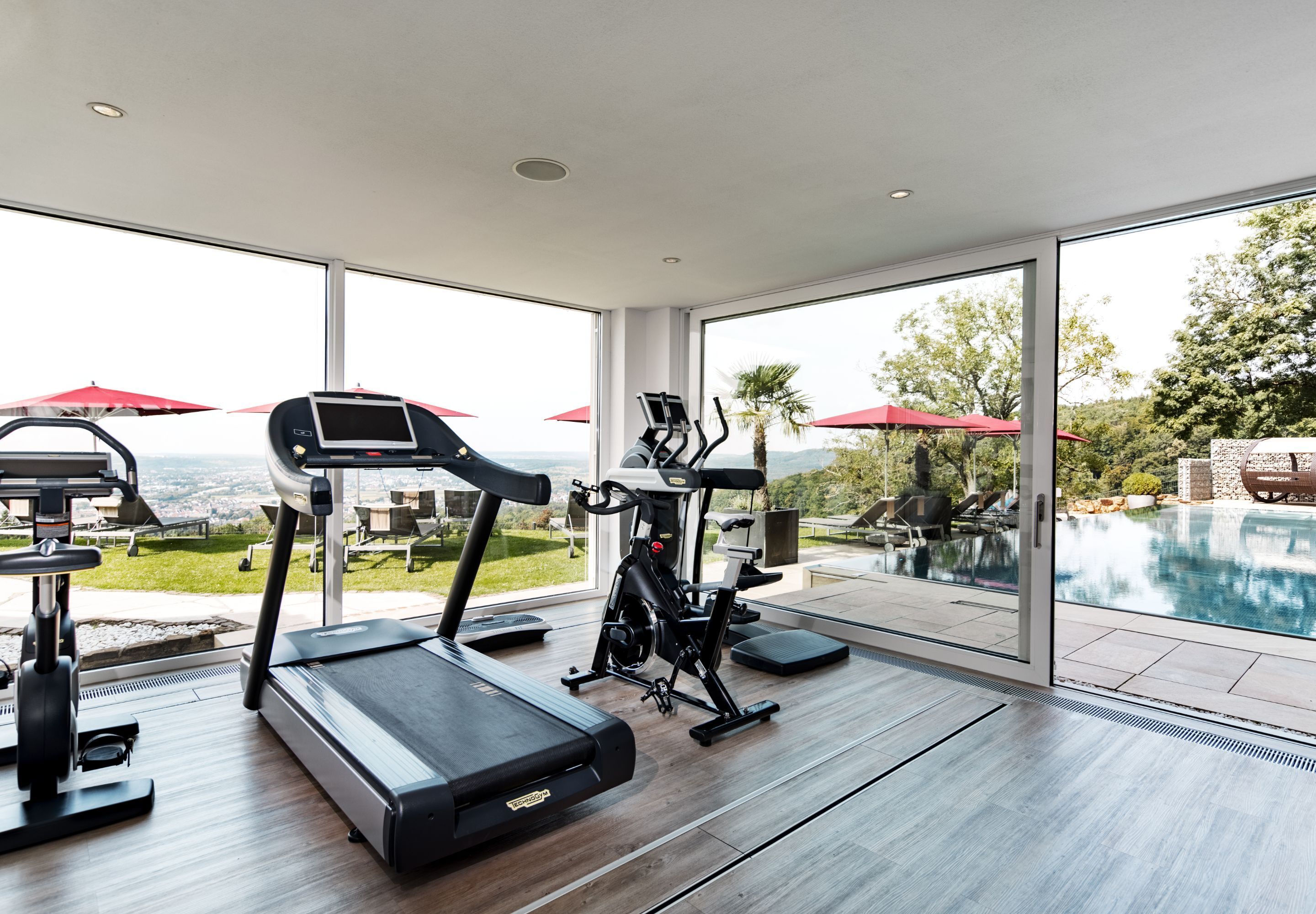 A modern gym with a treadmill, elliptical, and stationary bike, overlooking a pool and outdoor seating area.