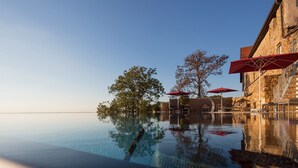 An infinity pool with clear water reflecting the sky and trees, surrounded by stone buildings and red umbrellas. - Burghotel Staufeneck (Salach)