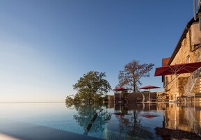Uma piscina infinita com água cristalina refletindo o céu e árvores, cercada por edifícios de pedra e guarda-sóis vermelhos.