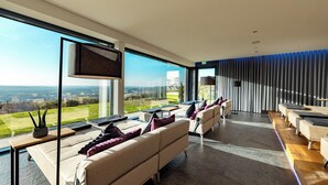 A modern hotel room with a large window offering a scenic view, a sofa with cushions, a flat-screen TV, and a dining area with a table and chairs. - Burghotel Staufeneck (Salach)